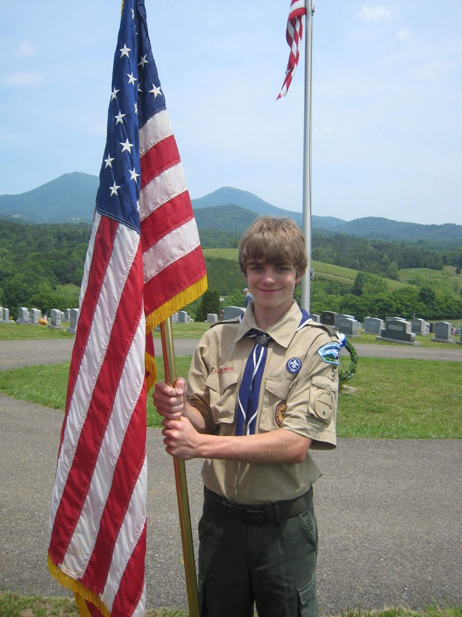 Ian de adolescente, con su uniforme y pañoleta de Boy Scout, sosteniendo una bandera estadounidense en un cementerio, con colinas verdes y montañas detrás.