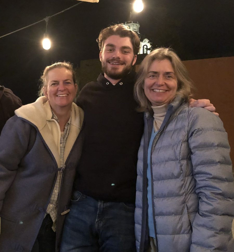 Ian al aire libre de noche, bajo guirnaldas de luces, de pie entre su madre Michelle y su tía Stephanie con un brazo alrededor de cada una, los tres sonriendo.