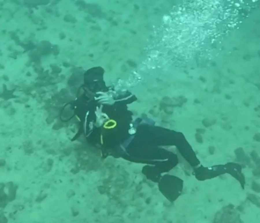 Ian buceando sobre un fondo marino arenoso y claro en agua verde transparente, con un rastro de burbujas subiendo desde su regulador.