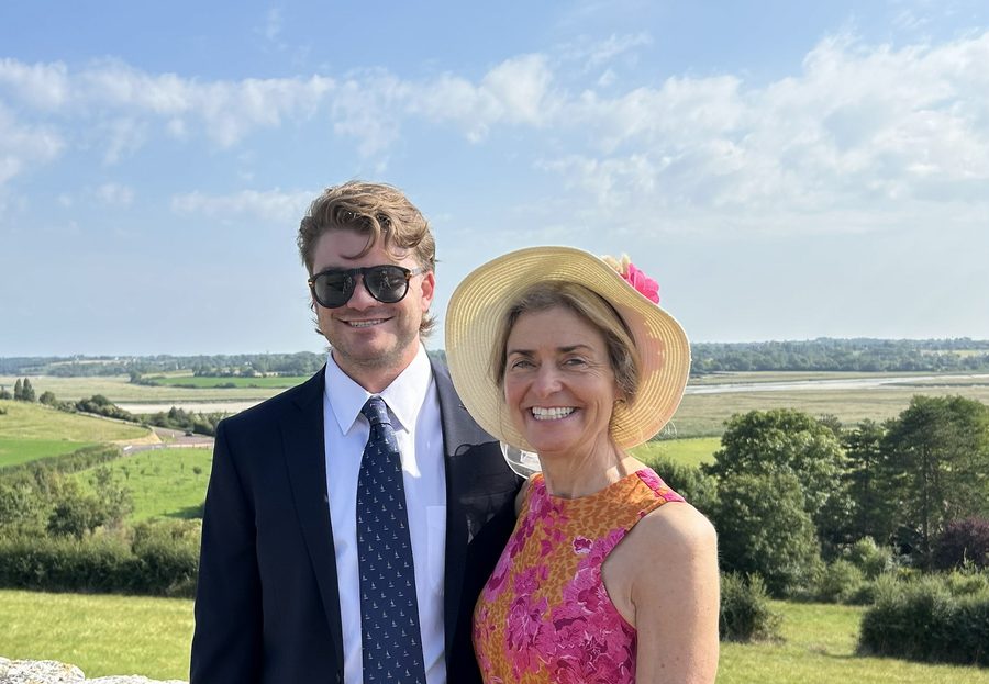 Ian con traje y corbata oscuros junto a su madre Michelle, con un sombrero de paja ancho y un vestido rosa y naranja, con el campo verde de Normandía y un estuario detrás.
