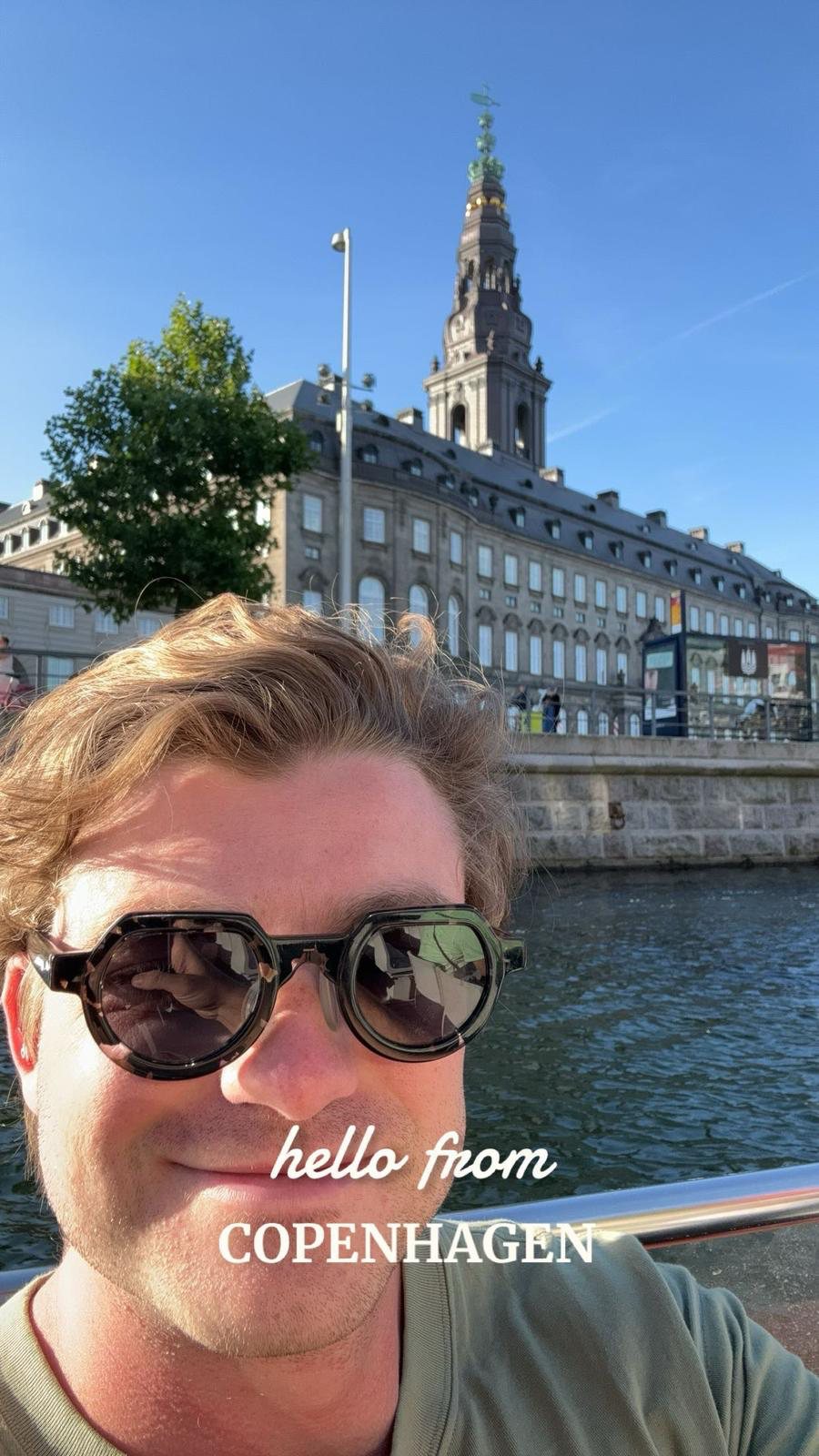 Ian con lentes de sol redondos, sonriendo a la cámara desde un bote en un canal de Copenhague, con la larga fachada y la aguja de techo verde del Palacio de Christiansborg detrás. Escrito sobre la fotografía, en inglés: hello from Copenhagen.