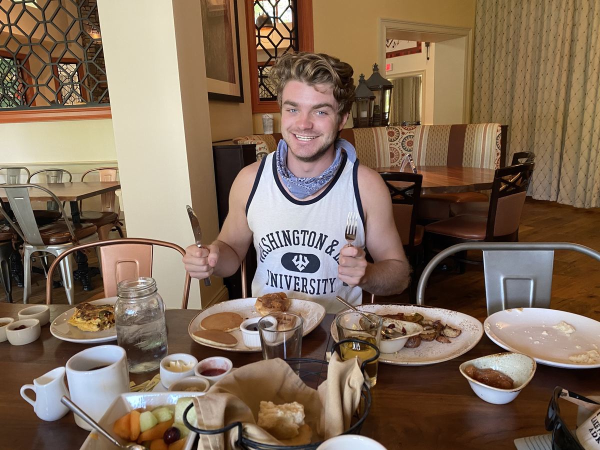 Ian sonriendo en una mesa de restaurante con un cuchillo en una mano y un tenedor en la otra, con una camiseta sin mangas de Washington and Lee University y una bandana, con la mesa frente a él cubierta de panqueques, bizcochos, papas, fruta y café.