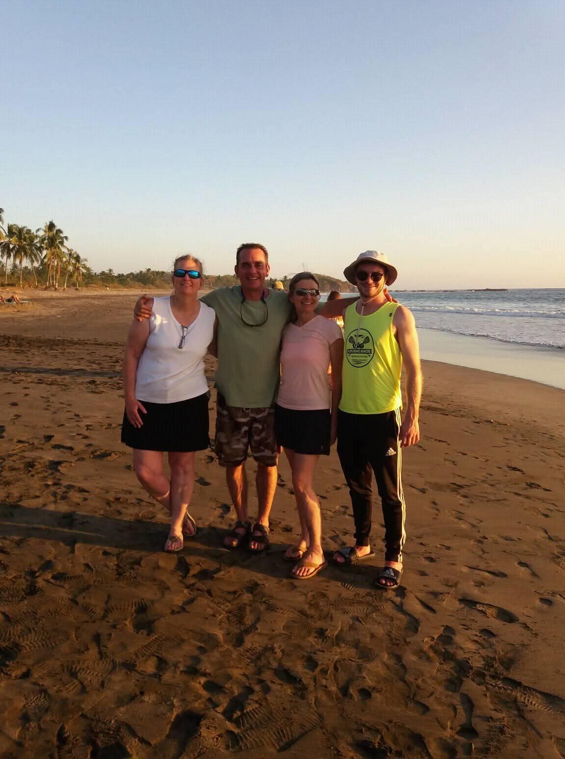 Ian con su madre, su tío Scott y su tía Stephanie, abrazados en una playa al atardecer, con palmeras y el oleaje detrás.