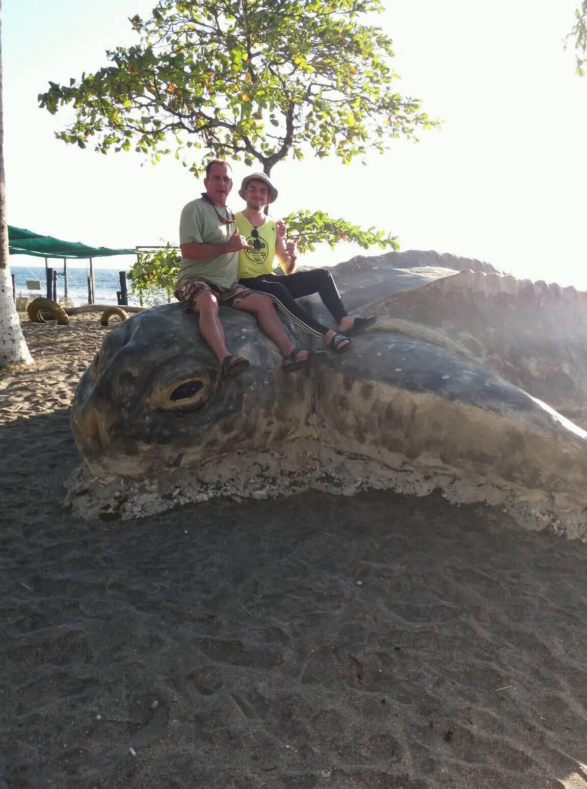 Ian y su tío Scott sentados uno al lado del otro sobre la cabeza de una gran estatua de tortuga marina bajo un árbol, ambos haciendo la señal shaka.