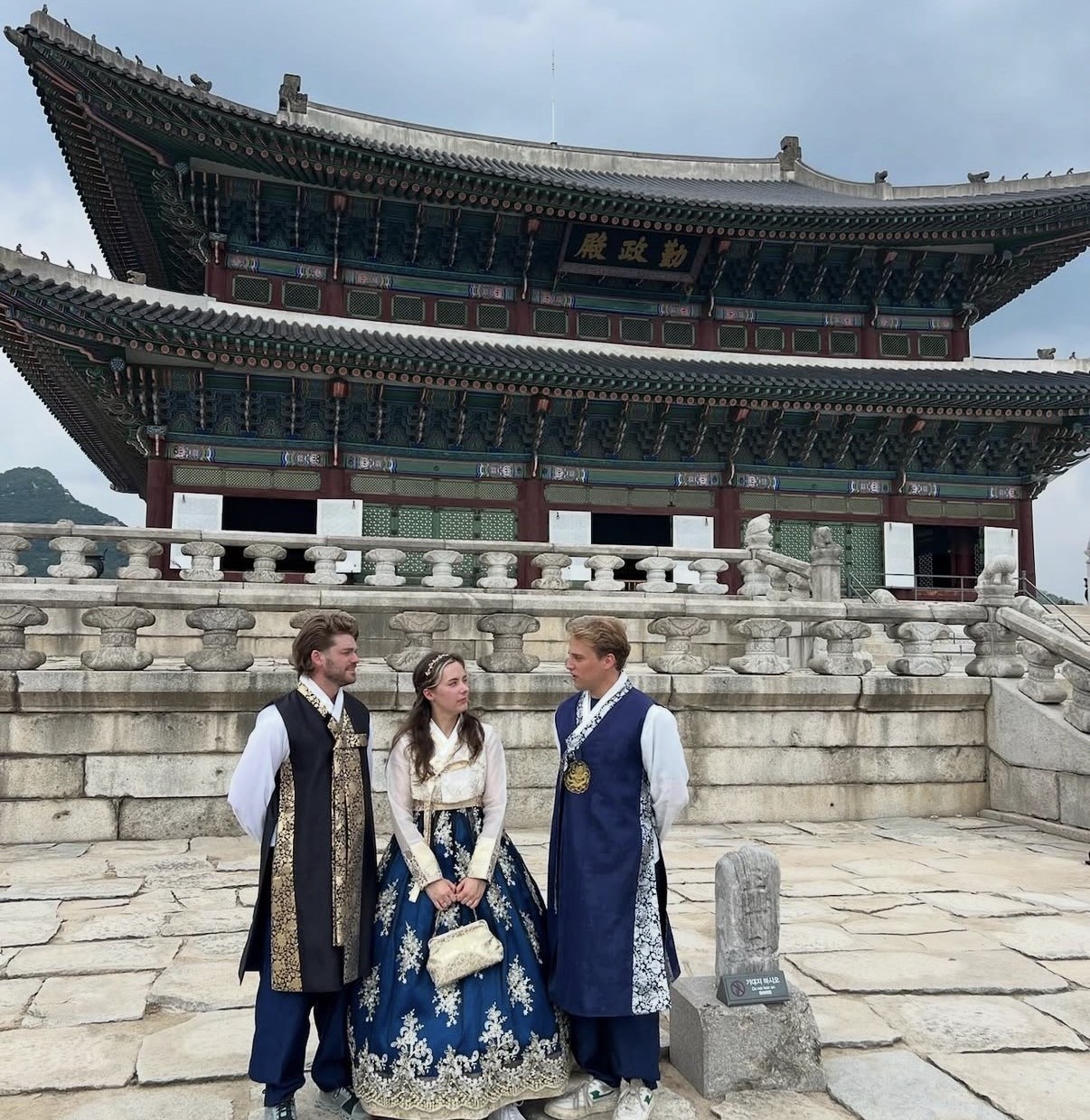 Ian y dos amigos vestidos con hanbok tradicional coreano, de pie frente al ornamentado salón Geunjeongjeon de techo verde en el Palacio Gyeongbokgung, Seúl.