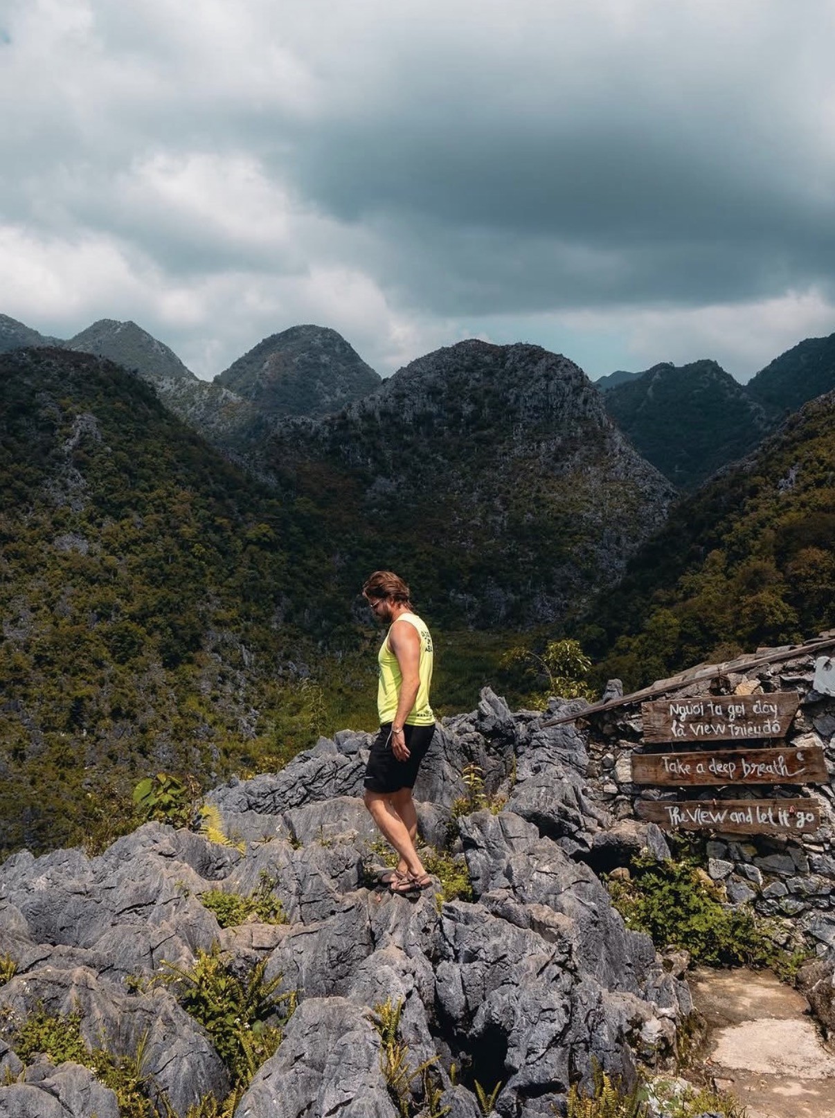 Ian, con una camiseta amarilla sin mangas, de pie sobre roca caliza irregular sobre un valle montañoso cubierto de neblina en el Ha Giang Loop, Vietnam, junto a un cartel de madera que dice, en inglés: “Take a deep breath, the view and let it go.”