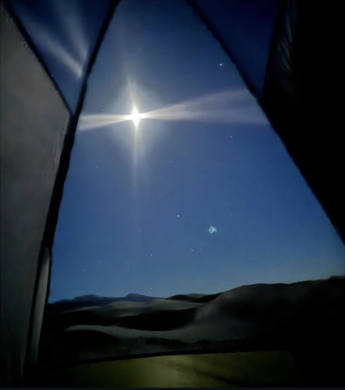 Una luna brillante con un destello en forma de cruz, y un tenue resplandor verdoso entre las estrellas, vista sobre una cresta montañosa oscura a través de la puerta abierta de una carpa.