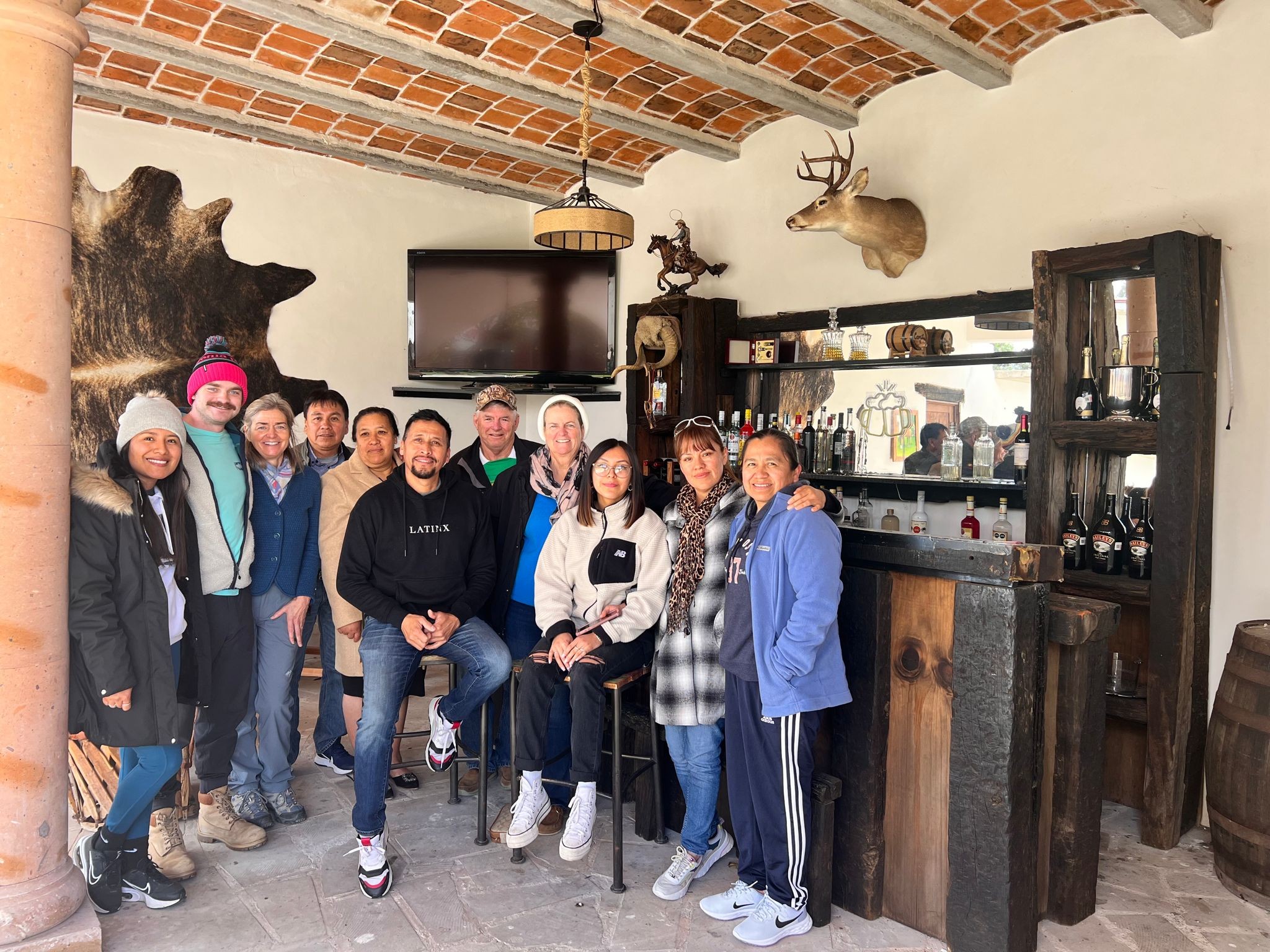 Ian, con un gorro rosa y una chaqueta verde azulado, reunido con cerca de una docena de familiares y amigos para una foto navideña en un bar rústico en México, con una cabeza de venado montada y un cuero de vaca en la pared detrás.