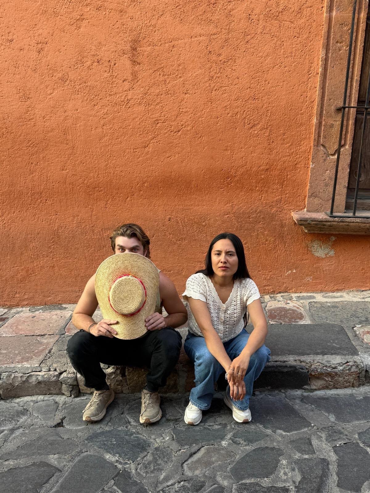 Ian, sentado en una acera contra una pared de color terracota, sosteniendo un sombrero de paja de ala ancha frente a su rostro, junto a su amiga Jazmin.