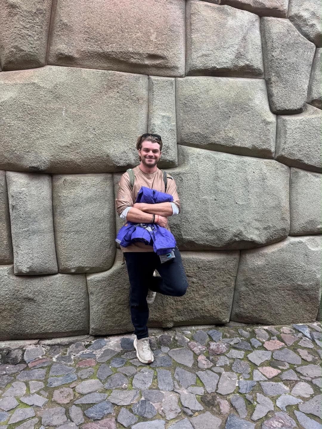 Ian sonriendo frente a un enorme muro de bloques de piedra inca perfectamente encajados, con una chaqueta doblada bajo el brazo.