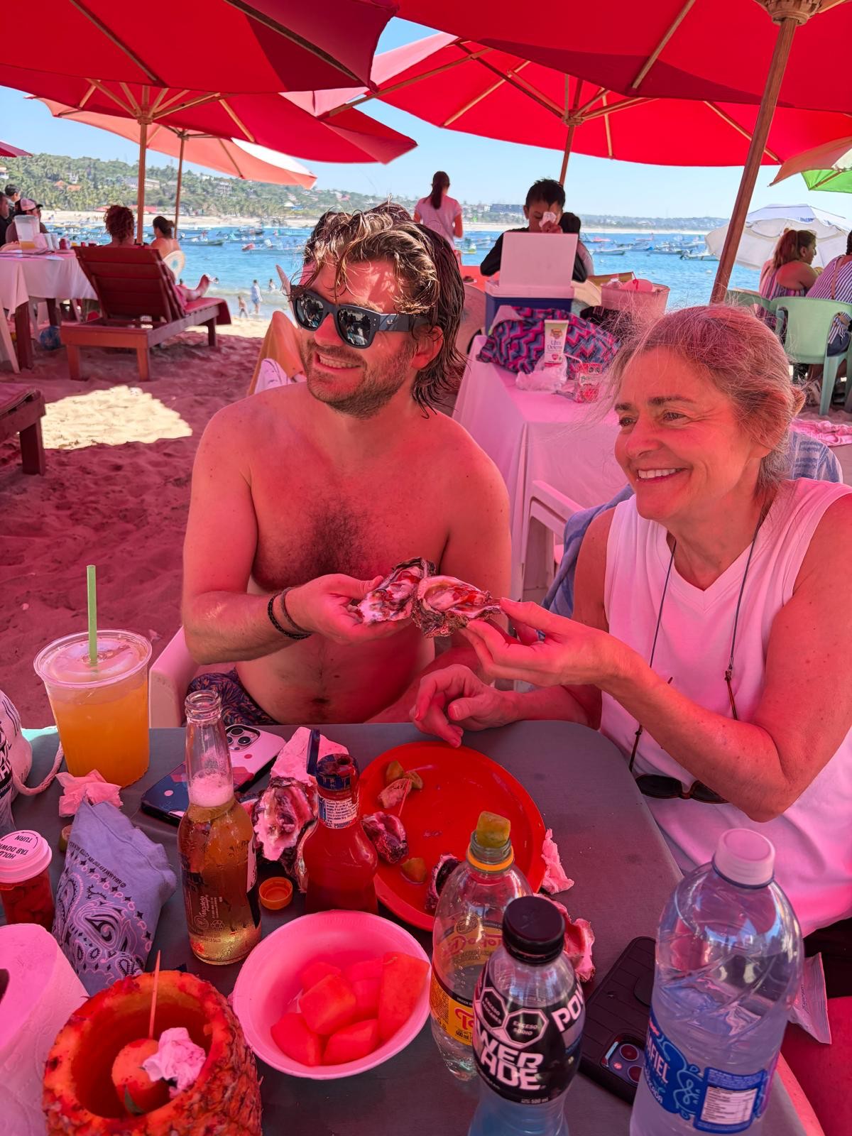 Ian, sin camisa y con lentes de sol, el pelo mojado, compartiendo ostras frescas con su madre Michelle en una mesa bajo sombrillas rojas de playa, con el mar detrás.