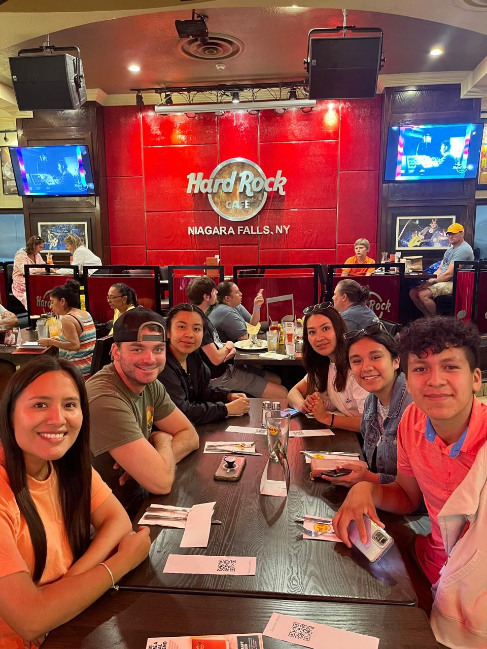 Ian, con gorra y camisa verde oliva, reunido en una mesa de restaurante con un grupo de amigos bajo un letrero iluminado que dice “Hard Rock Cafe, Niagara Falls, NY”.