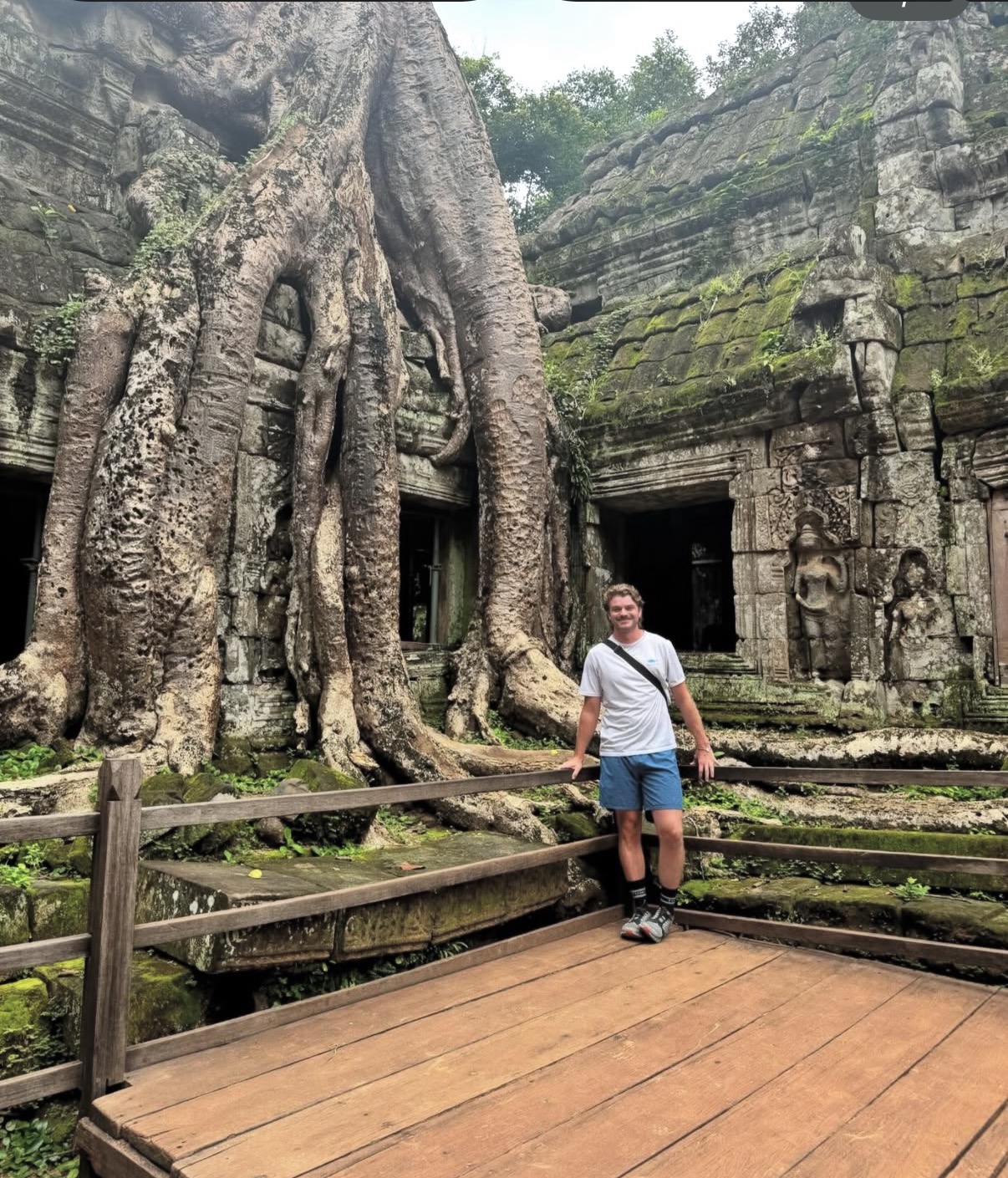 Ian de pie sobre una pasarela de madera en las ruinas del templo de Ta Prohm, Camboya, empequeñecido por enormes raíces de árboles que envuelven las antiguas torres de piedra junto a una figura tallada en piedra.