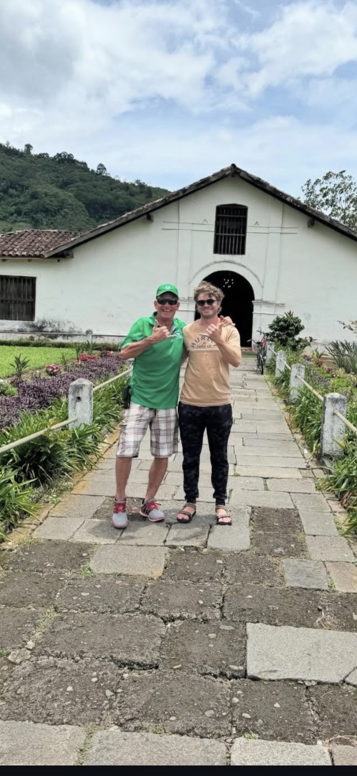 Ian y su tío Scott, ambos haciendo la señal shaka, de pie del brazo sobre un camino de piedra frente a un edificio blanco de estilo colonial con techo de tejas, con montañas verdes detrás.