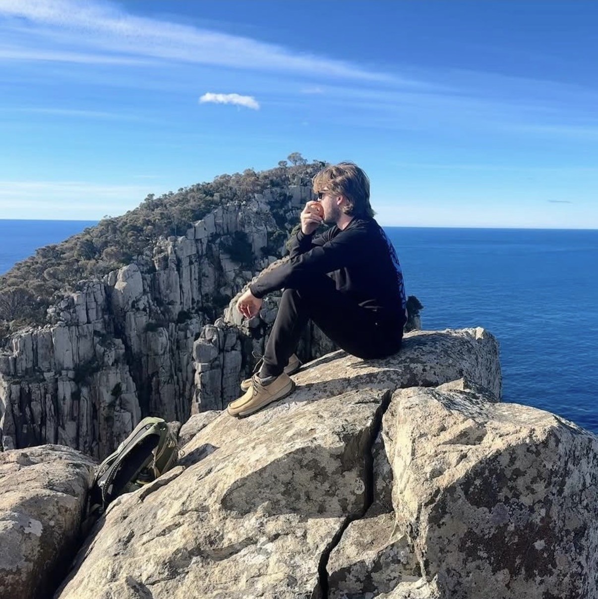 Ian sentado con las piernas cruzadas al borde de un acantilado rocoso con vista al mar y a una cresta montañosa en la cordillera Western Arthur, Tasmania, comiendo una manzana.
