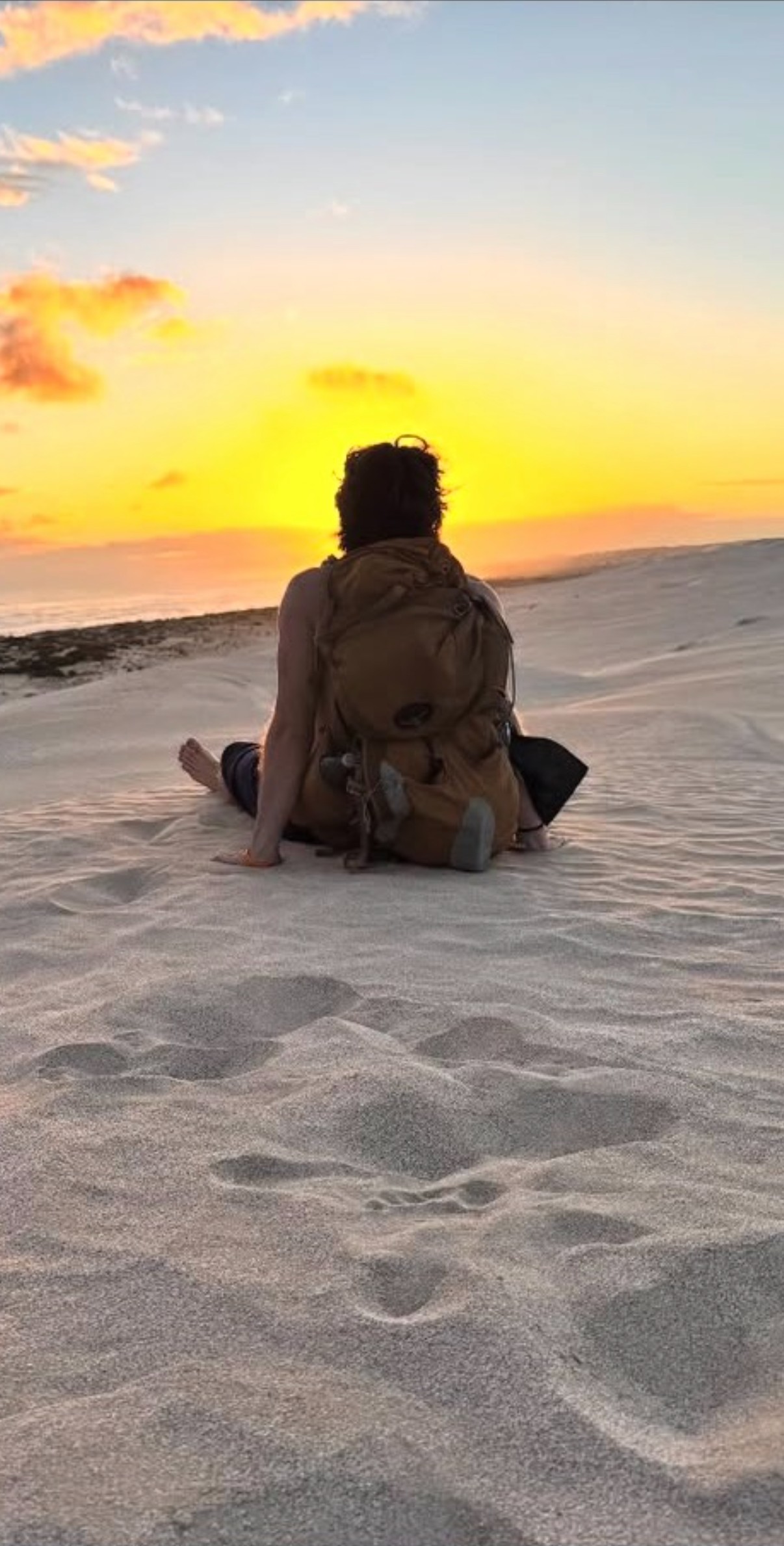 Ian, visto de espaldas y con una mochila de excursionismo, sentado en una duna de arena mirando un vívido atardecer naranja y amarillo sobre el mar en Australia.