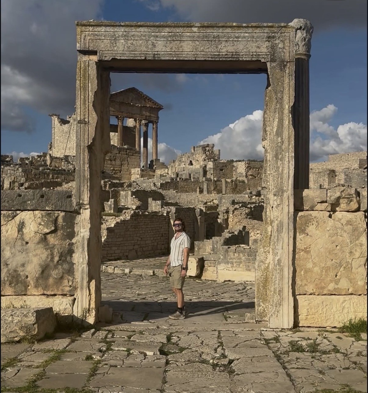 Ian de pie bajo un arco de piedra en las antiguas ruinas romanas de Dougga (Thugga), en Túnez, con un templo de columnas visible al fondo.
