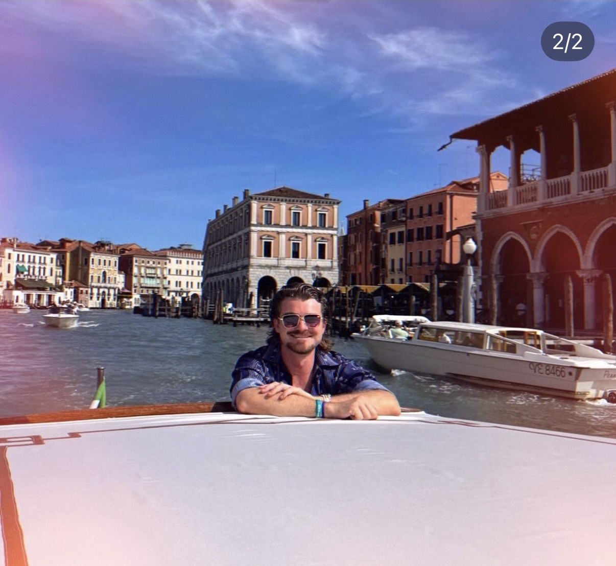 Ian, con lentes de sol, apoyado en la baranda de un bote en el Gran Canal de Venecia, con edificios históricos frente al agua y otros botes detrás.