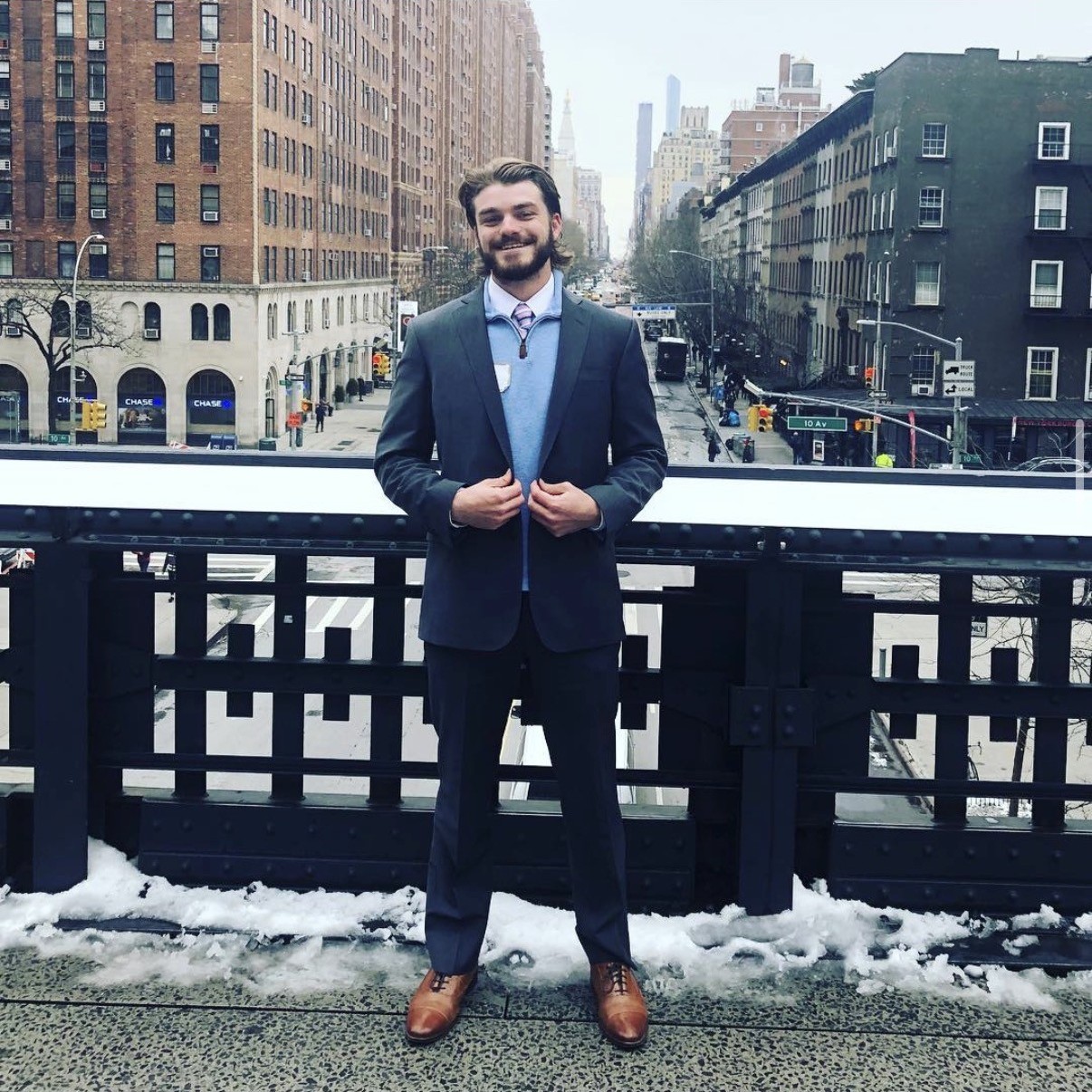 Ian, con traje y corbata, sonriendo en el High Line de Nueva York con el horizonte de Manhattan y una baranda cubierta de nieve detrás.