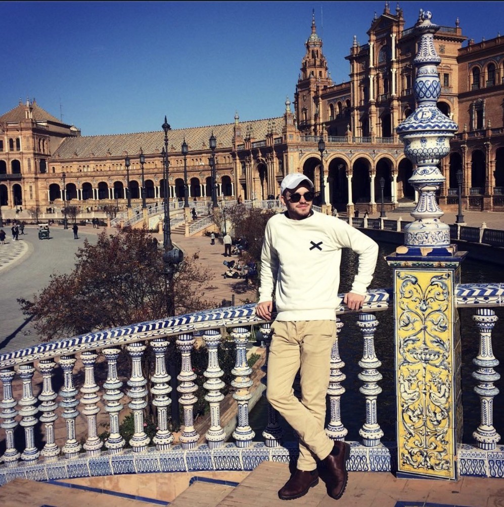 Ian, con gorra blanca y lentes de sol, apoyado contra una balaustrada de azulejos ornamentados sobre el foso de la Plaza de España en Sevilla, España.