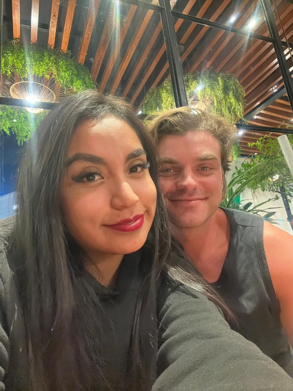 Ian y su amiga Silvana tomándose una selfie sonriendo, bajo un techo de madera cubierto de plantas.