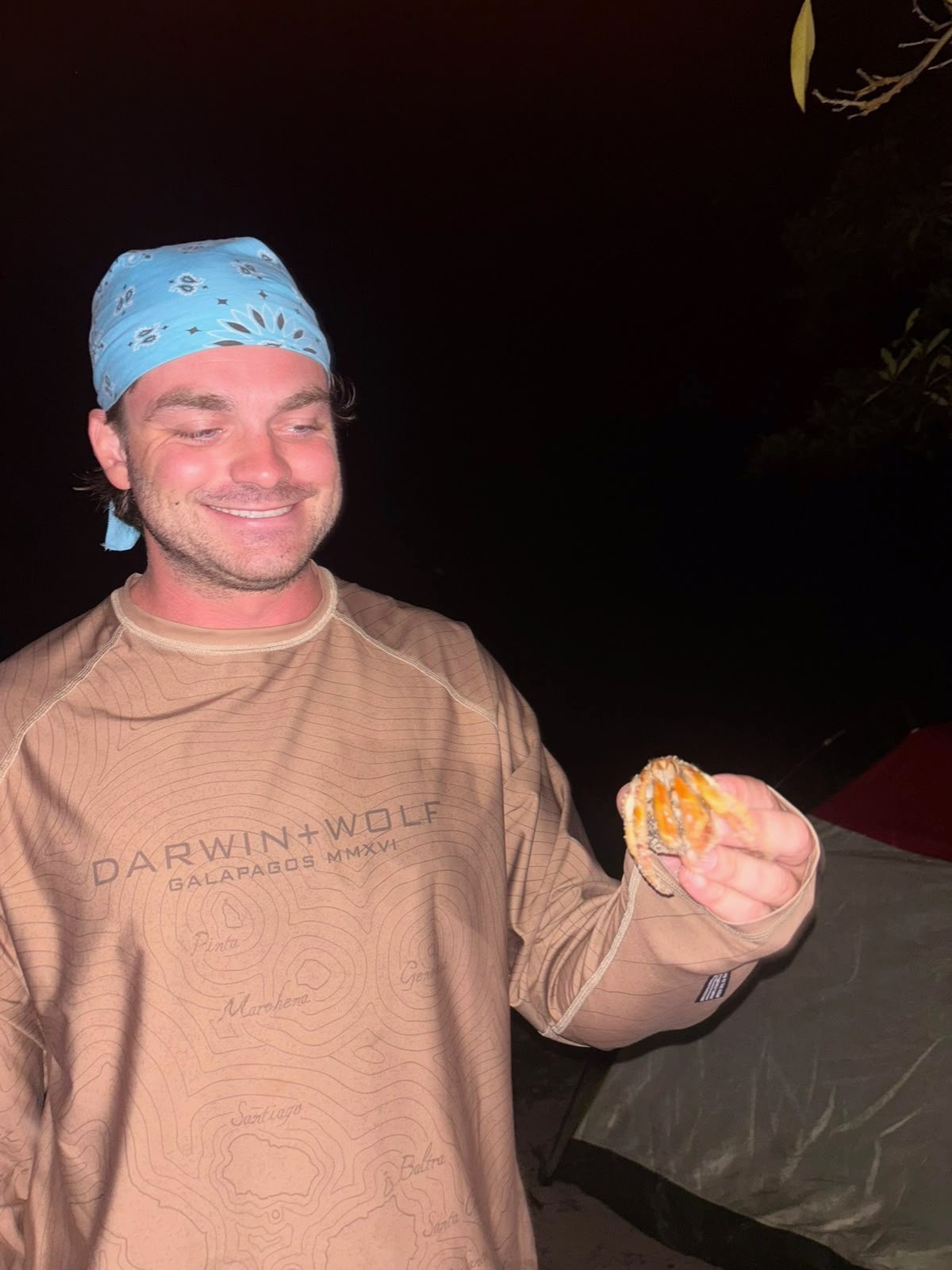 Ian, con una bandana azul y una camisa estampada con un mapa de las Islas Galápagos, sonriendo de noche mientras sostiene un pequeño cangrejo para mirarlo.