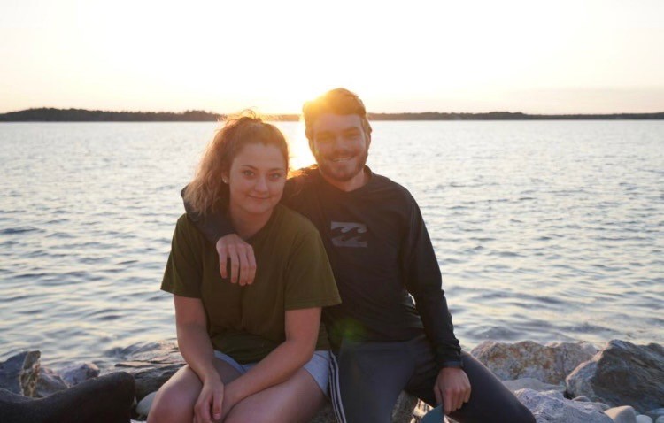 Ian y su amiga Marisa sentados juntos sobre rocas a la orilla del agua en Nueva Escocia al atardecer, con la mano de ella apoyada en el hombro de él.