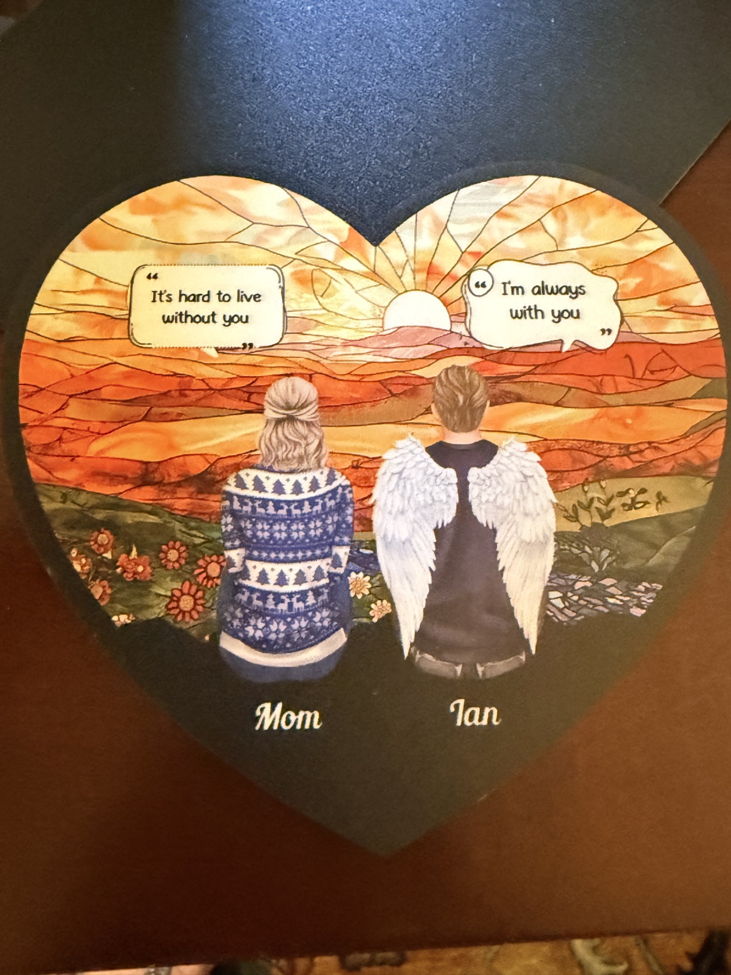 Un recuerdo conmemorativo ilustrado: Ian, con alas de ángel, y su madre sentados juntos mirando un atardecer, con las palabras, en inglés, “It's hard to live without you” y “I'm always with you” junto a ellos.
