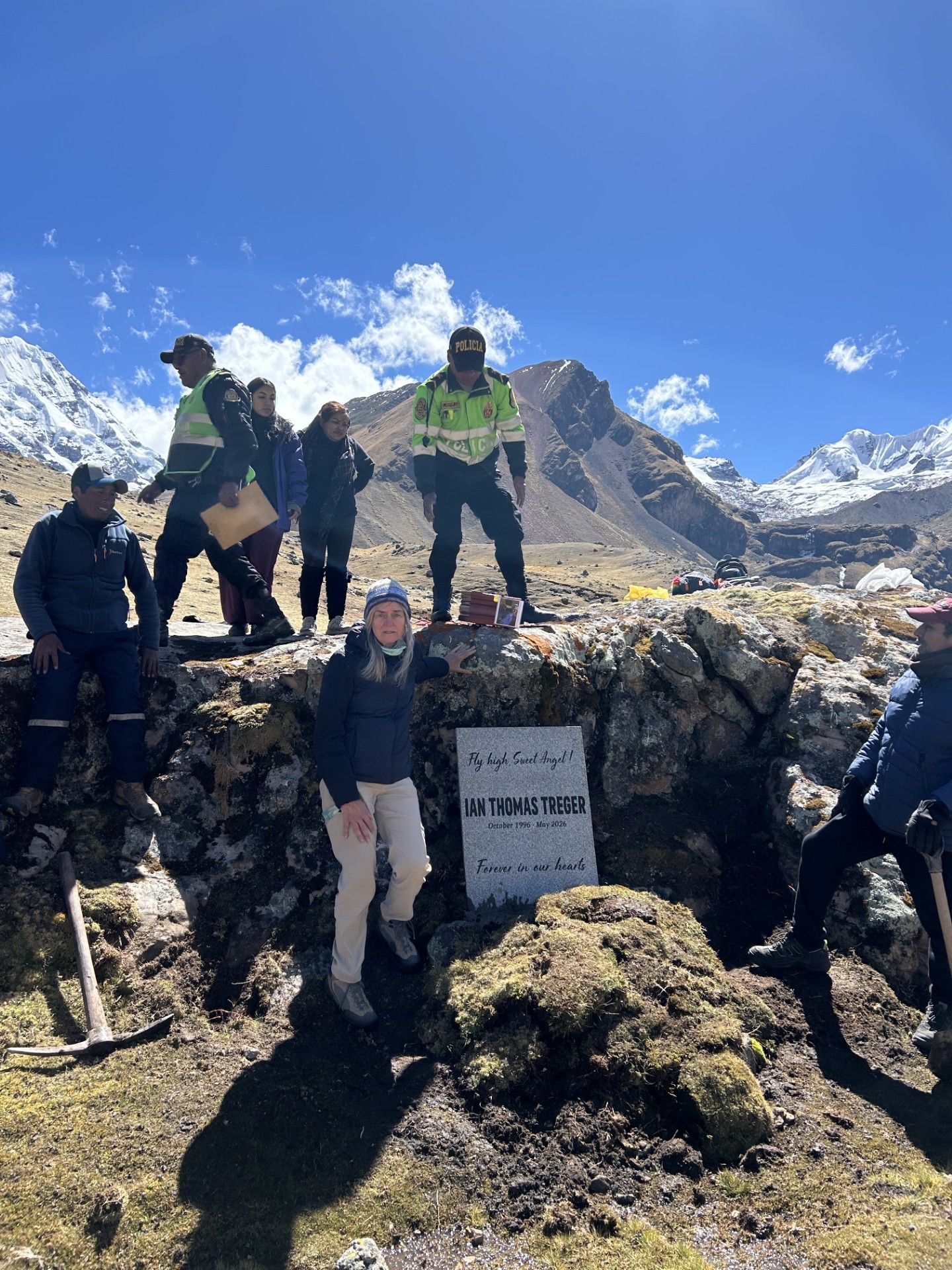 La madre de Ian, Michelle, junto a una piedra conmemorativa que dice, en inglés, “Fly high, Sweet Angel! Ian Thomas Treger, October 1996 – May 2026, Forever in our hearts,” colocada entre rocas altas en los Andes peruanos, rodeada de policías peruanos y amigos que ayudaron a cargar la piedra con las cenizas de Ian hasta este lugar espiritual, con montañas nevadas detrás.