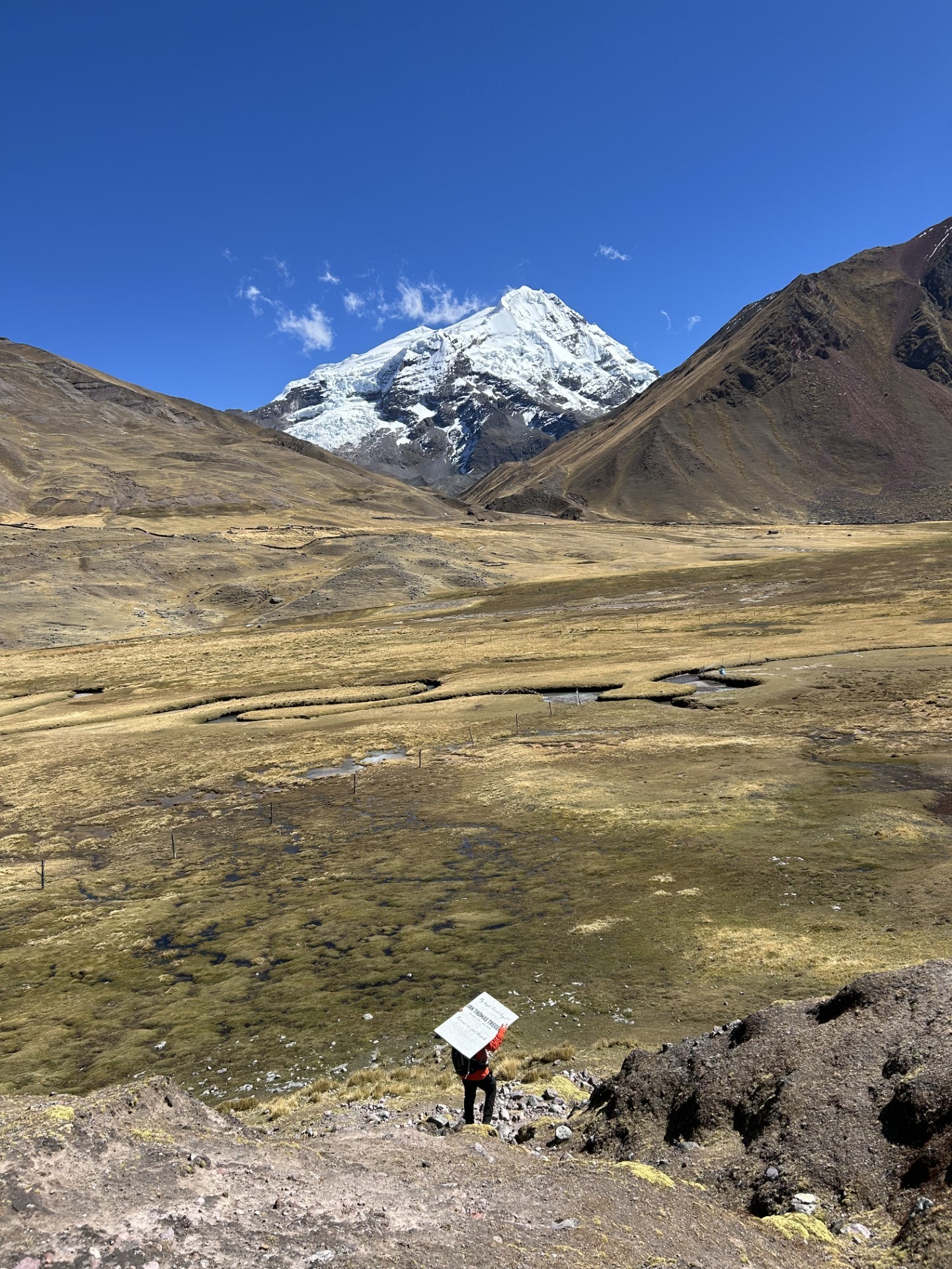 Henrry, visto a la distancia, cargando la piedra conmemorativa de Ian sobre su cabeza a través de un vasto valle andino en Perú, con un pico nevado al fondo.
