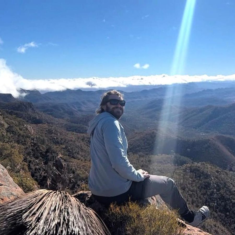 Ian Treger sentado en una cumbre rocosa, con lentes de sol, mirando hacia la cámara sobre un valle y un manto de nubes.