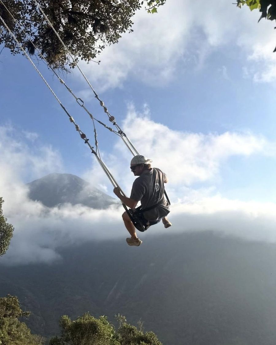 Ian Treger en un columpio de cuerda sobre un valle cubierto de nubes, con una montaña asomando entre las nubes detrás de él.