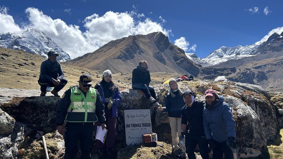 Ian's memorial marker on Ausangate, snow-capped peaks behind it, with a small group of family and guides gathered around. The marker reads: Fly high, Sweet Angel! Ian Thomas Treger, October 1996 – May 2026, Forever in our hearts.