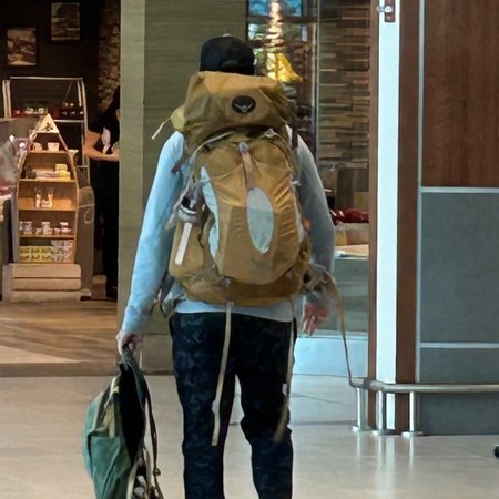 Ian caminando por una terminal de aeropuerto, con una mochila de excursionismo completa y cargando un bolso.